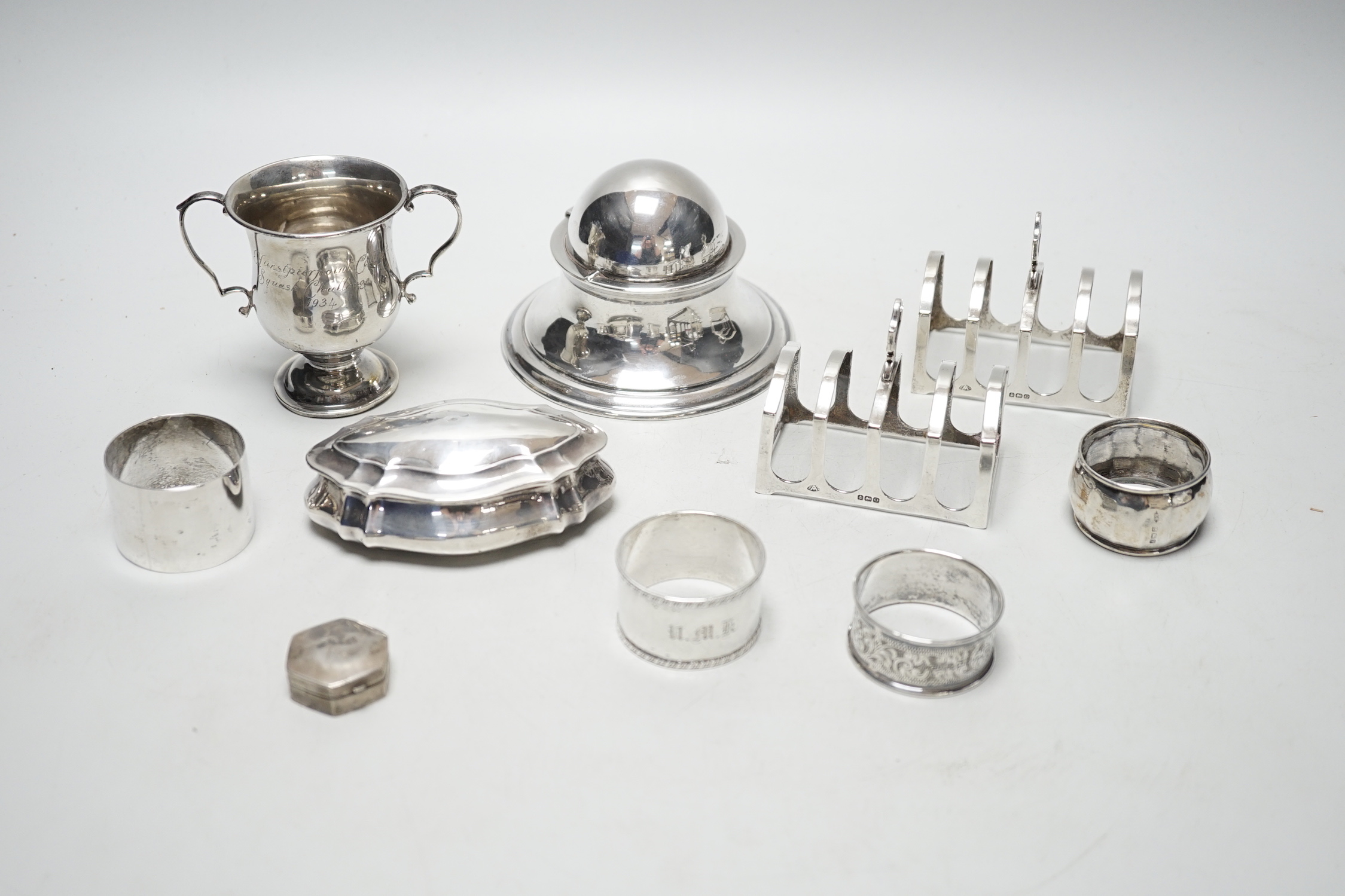 A late Victorian shaped oval silver trinket box, Birmingham, 1898, 99mm, a hexagonal silver pill box, a pair of silver toast racks and four silver serviette rings, a silver domed topped inkwell and a miniature silver two